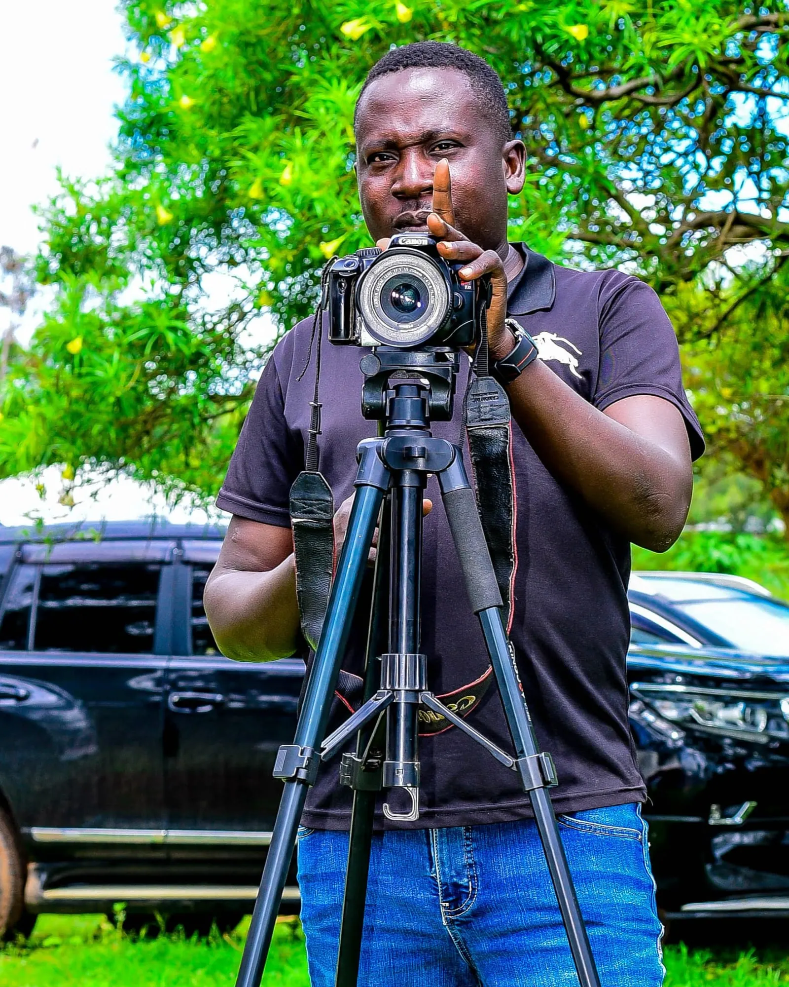 A cameraman behind a camera on a tripod, filming outdoors under a tree